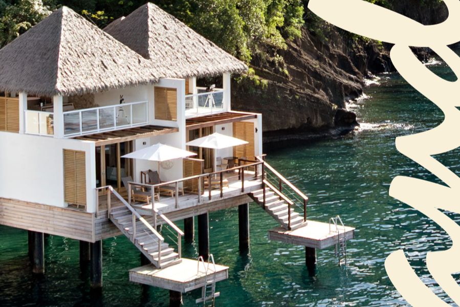 A modern stilted villa with a thatched roof overlooks calm turquoise waters, featuring wooden steps leading to a small platform, surrounded by lush greenery and rocky cliffs, featured in the Zola Sandals Sweepstakes