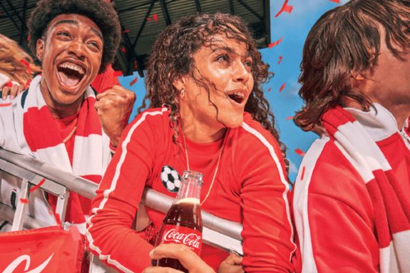 Excited fans celebrate at a sports event, wearing red apparel. One woman joyfully holds a drink while others cheer, surrounded by festive red confetti and a vibrant atmosphere, featured in the Coca-Cola FIFA World Cup Sweepstakes