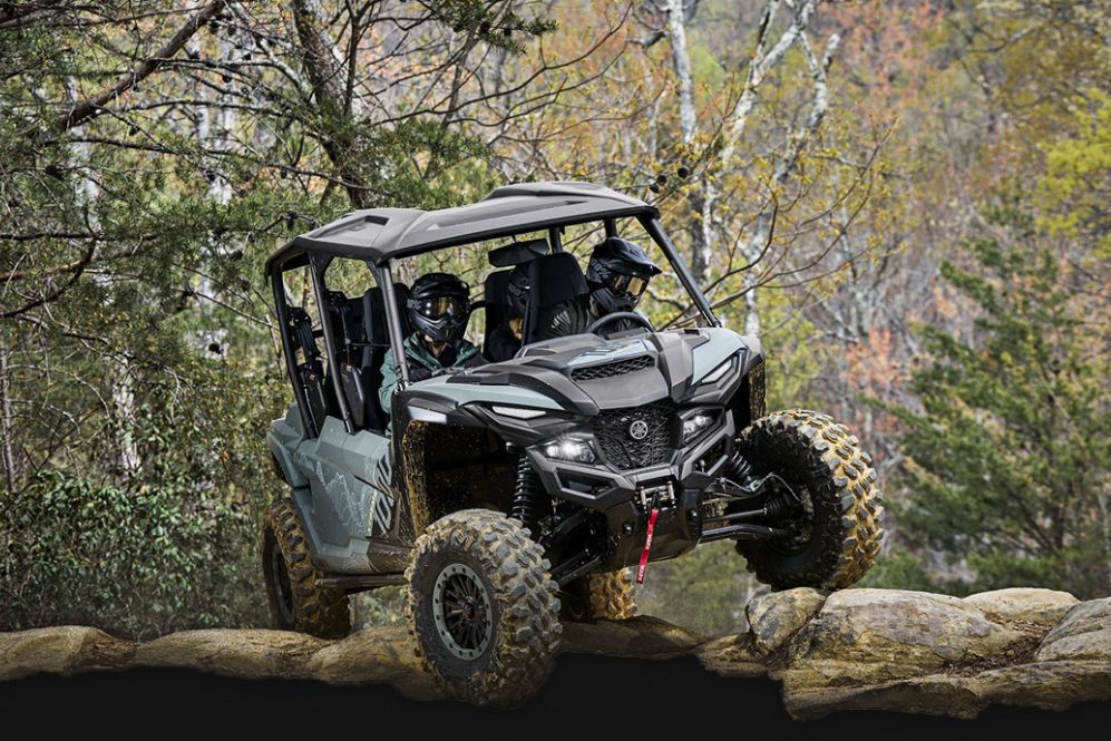 A powerful all-terrain vehicle navigates over rocks in a forested area, with two helmeted riders seated inside, showcasing off-road capabilities against a backdrop of trees, featured in the PenFed Yamaha Wolverine Sweepstakes