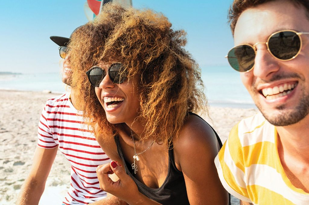 Three friends laugh together on a sunny beach, enjoying the warm weather. One has curly hair and sunglasses, while the others wear casual beach attire and sunglasses, radiating joy, featured in the Barefoot Here Comes The Fun Sweepstakes