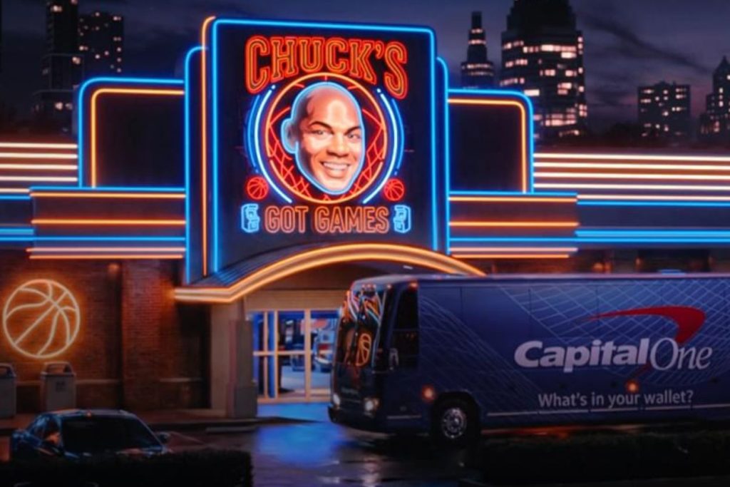 A brightly lit building features a large neon sign that displays "Chuck's Got Games," while a blue truck is parked nearby, set against a city skyline at dusk, featured in the Capital One March Madness Sweepstakes