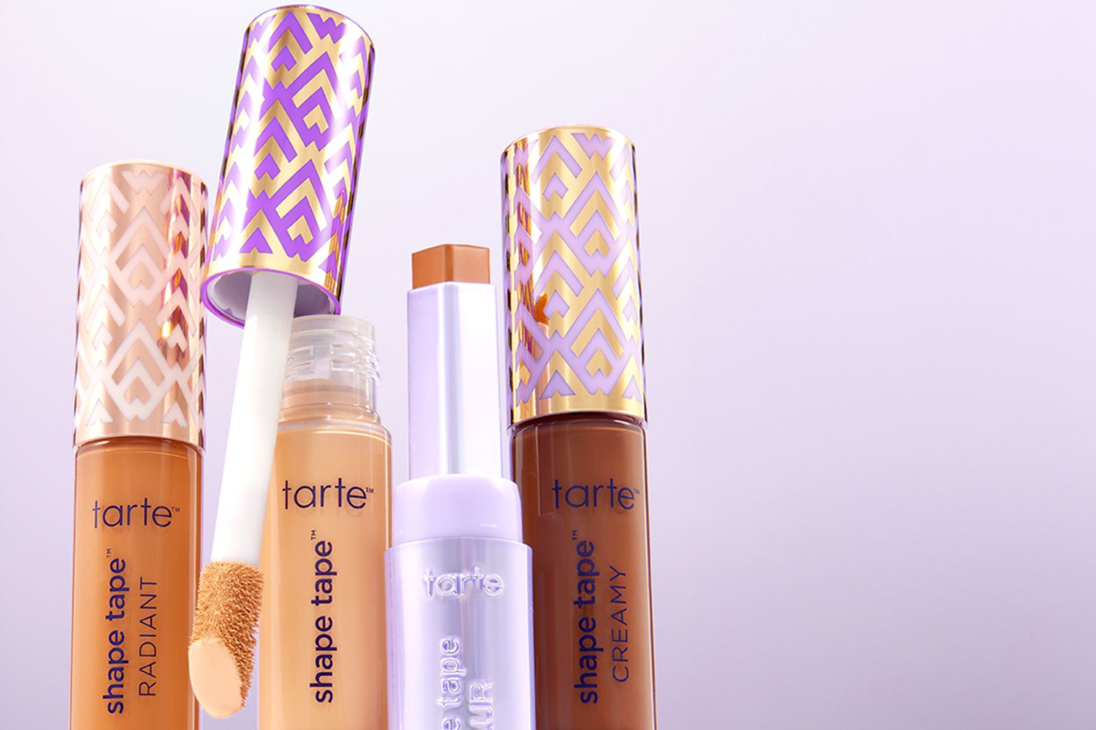 Four makeup products are displayed against a light purple backdrop, showcasing concealers and a contour stick with ornate packaging. The products feature various shades for skin tones, featured in the Tarte Shape Tape for a Year Sweepstakes
