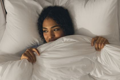 A woman with curly hair smiles while snuggled under a cozy white comforter in a softly lit bedroom, exuding a sense of warmth and comfort, featured in the World Sleep Day Sweepstakes