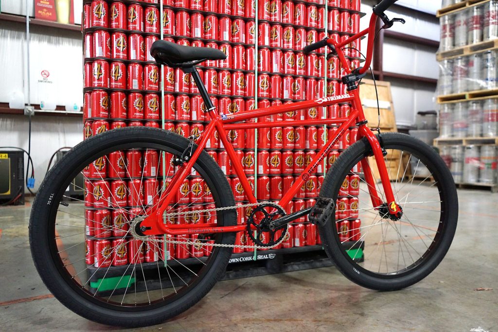 A bright red bicycle stands prominently in front of a towering stack of red cans, creating a striking contrast in a warehouse setting. The bike features a classic design with thick tires, featured in the Real Ale Brewing Cruzer Bicycle Sweepstakes