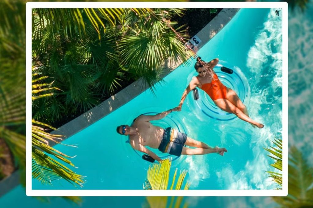 A couple floats on inner tubes in a turquoise lazy river, smiling and holding hands, surrounded by lush palm leaves under bright sunlight, featured in the Honeyfund Spring Getaway Sweepstakes