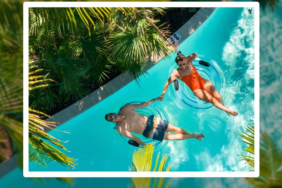 A couple floats on inner tubes in a turquoise lazy river, smiling and holding hands, surrounded by lush palm leaves under bright sunlight, featured in the Honeyfund Spring Getaway Sweepstakes