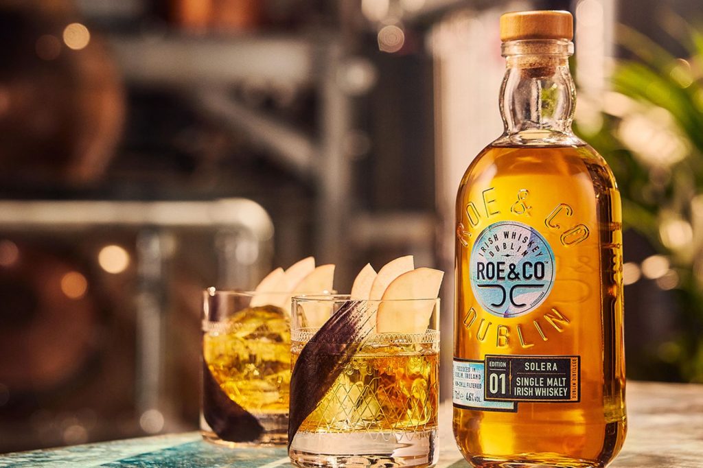 A bottle of Irish whiskey sits beside two glasses filled with golden whiskey, garnished with thin apple slices, against a softly blurred background of distillation equipment, featured in the GUINNESS ST PATRICKS DAY IRELAND SWEEPSTAKES