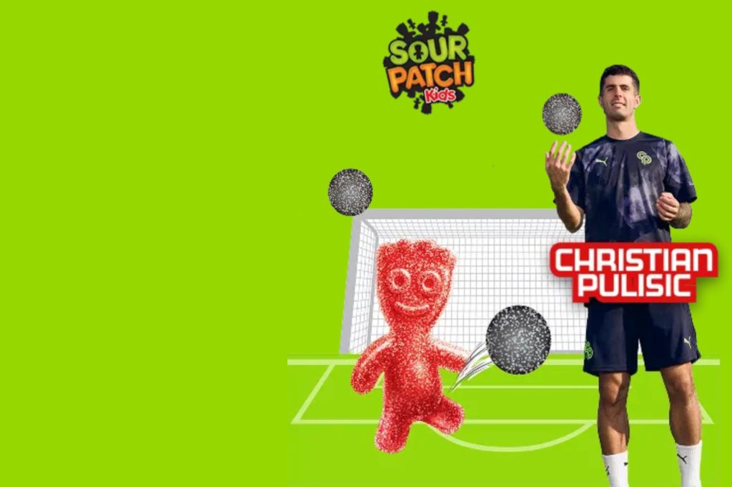 A soccer player juggles while a playful red gummy character runs on a bright green background near a goalpost, adding a fun, energetic vibe to the scene, featured in the Sour Patch Kids Soccer Jersey Sweepstakes