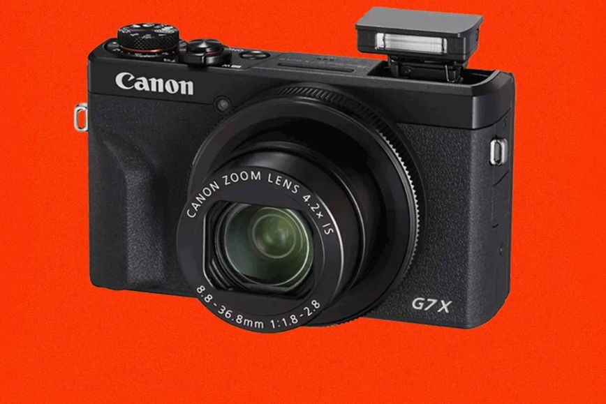 A sleek black camera with a zoom lens is displayed against a vibrant orange background, highlighting its compact design and professional features, featured in the P.E Nation Canon PowerShot G7X Sweepstakes