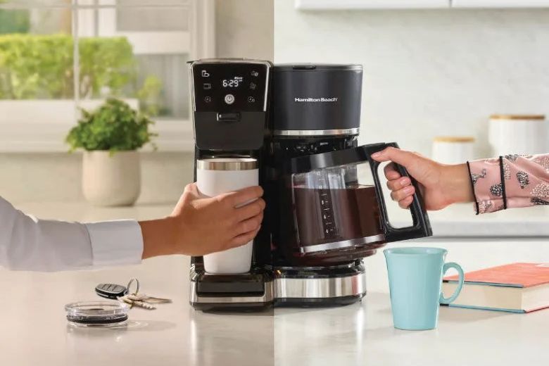 A person pours coffee from a machine into a metal travel mug while another hand reaches for a glass coffee pot. A light, bright kitchen setting enhances the scene, featured in the Hamilton Beach 2-Way Coffee Maker Sweepstakes