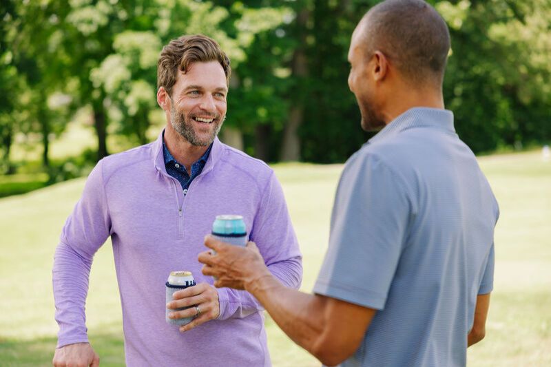 Two men enjoy a friendly conversation outdoors, holding cans in their hands. One man is smiling, wearing a light purple pullover, while the other is dressed in a gray polo shirt, featured in the Turtleson $1,000 Shopping Spree Sweepstakes