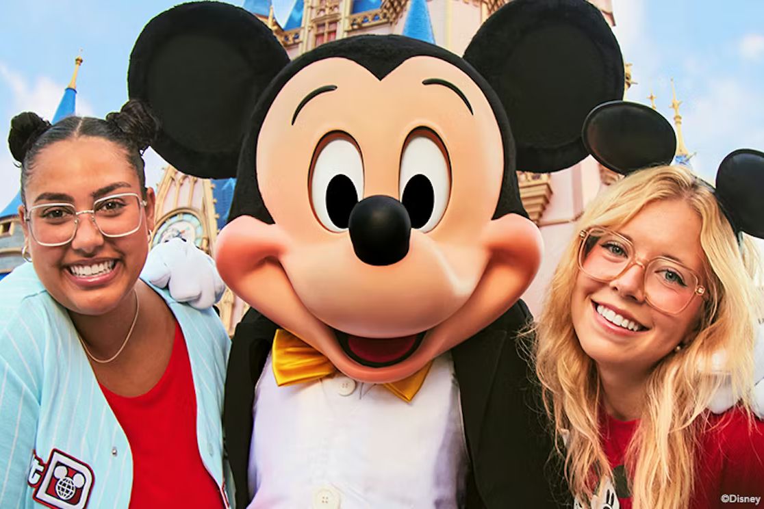 Two smiling young women pose with a large cartoon character wearing a tuxedo and bowtie, set against a whimsical castle backdrop with blue spires, featured in the BDG Disney World Sweepstakes