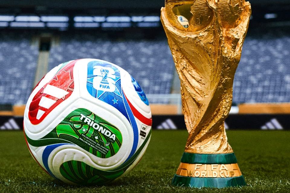 A colorful soccer ball rests on the green field next to a gleaming FIFA World Cup trophy, set against the backdrop of a stadium filled with empty seats, featured in the adidas FIFA World Cup Final Sweepstakes