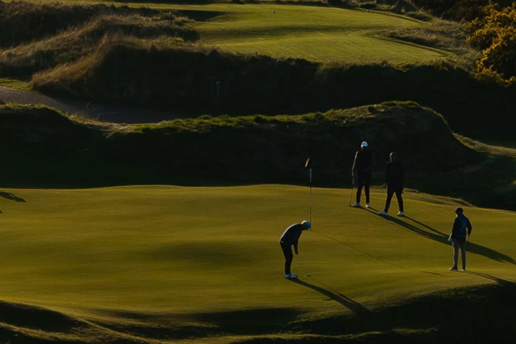 A golfer lines up a putt on a lush green course while three others observe. The setting sun casts long shadows across the landscape, creating a serene atmosphere, featured in the TravisMathew St Andrews Trip Sweepstakes