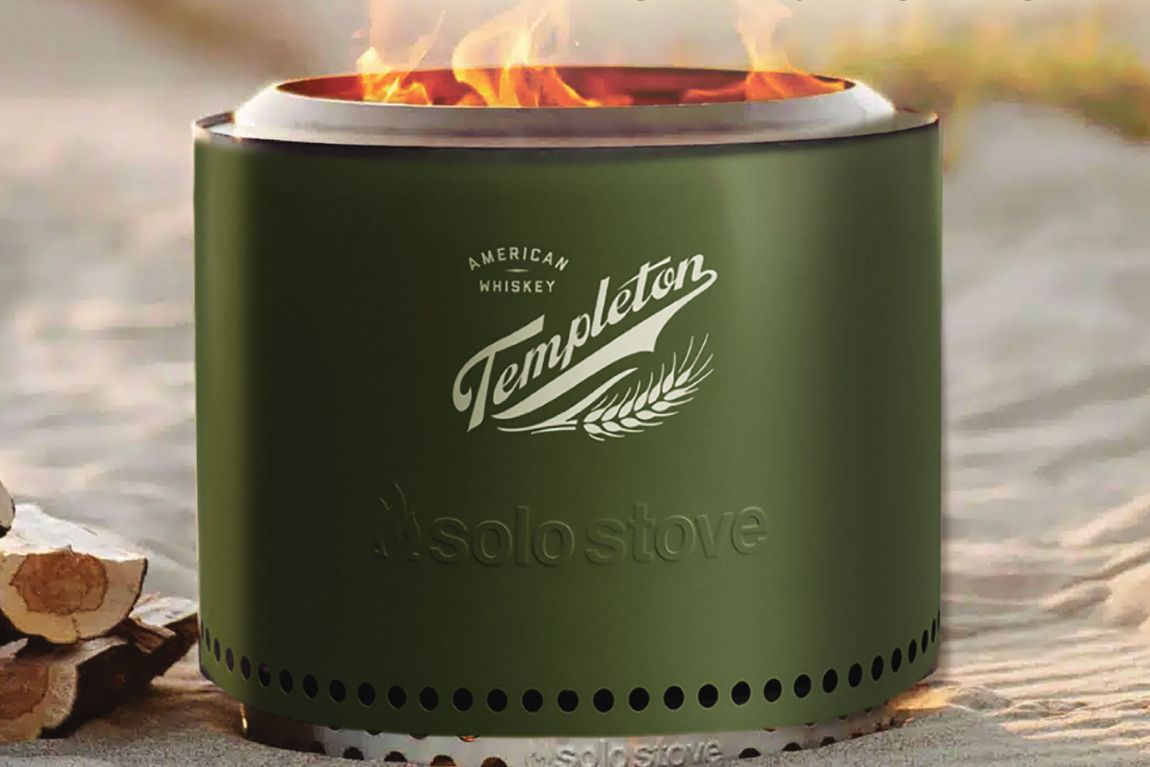 A sleek green portable fire pit sits on sandy terrain, with flames flickering from the top, surrounded by stacked logs, creating a cozy outdoor atmosphere, featured in the Templeton Whiskey Backyard Upgrade Sweepstakes