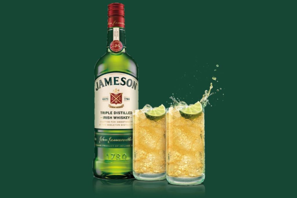 A bottle of Irish whiskey stands beside two glasses filled with a sparkling drink and ice, with lime wedges adding garnish, against a smooth green backdrop, featured in the Jameson St. Patrick's Day Sweepstakes