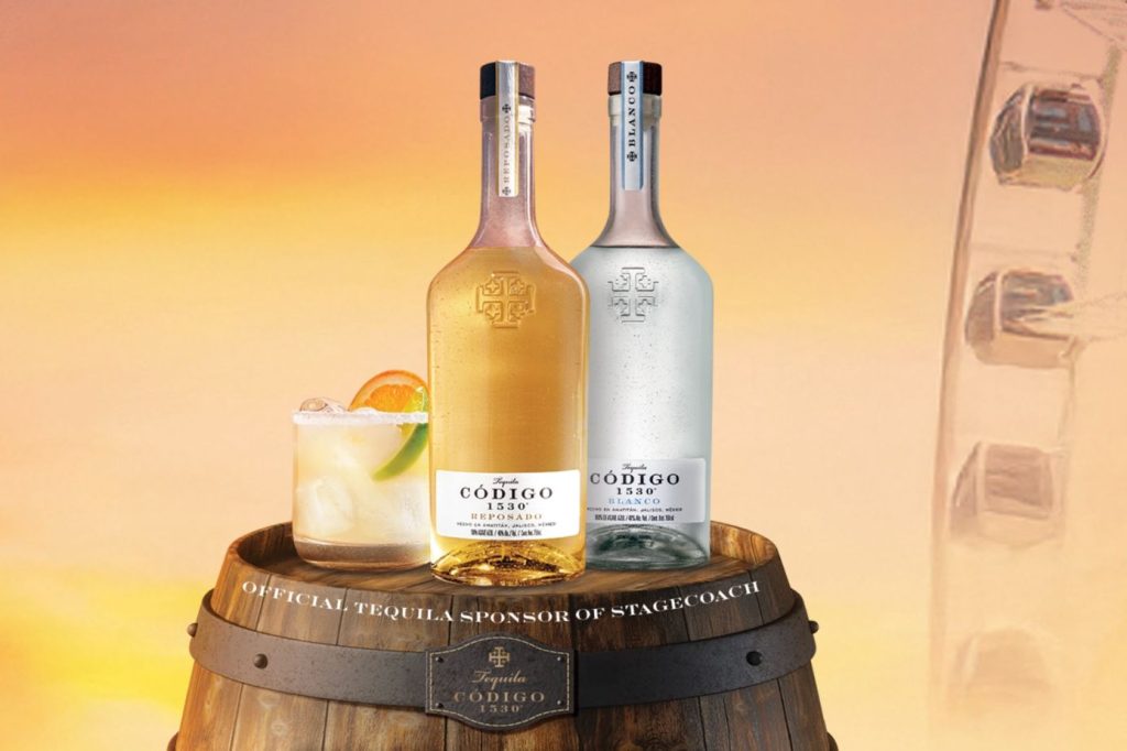 Two bottles of Código 1530 tequila, one reposado and one blanco, are displayed on a wooden barrel beside a refreshing cocktail garnished with fruit, set against a warm, sunset backdrop, featured in the Codigo 1530 Tequila Stagecoach Sweepstakes