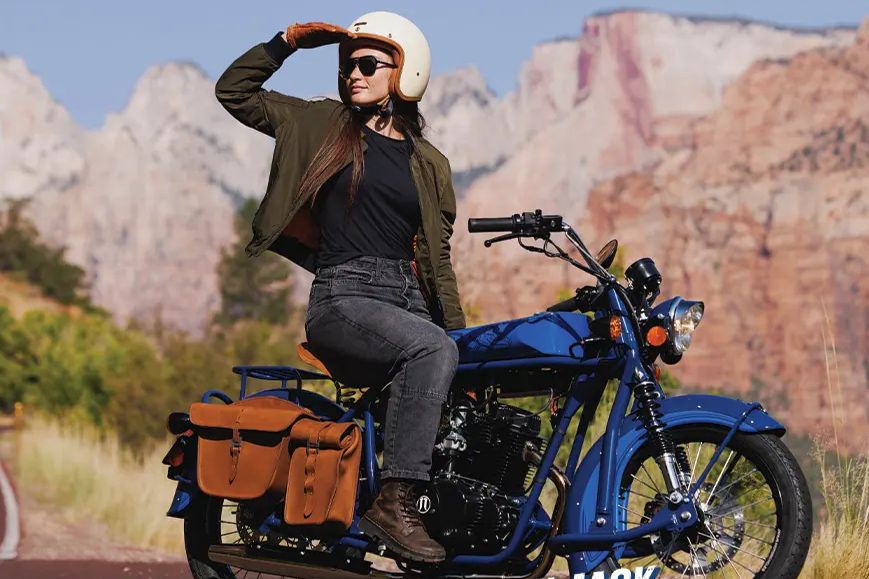 A woman wearing a helmet and sunglasses sits confidently on a blue motorcycle, shielding her eyes from the sun, with rugged mountains in the background, featured in the Cayman Jack Motorcycle Sweepstakes