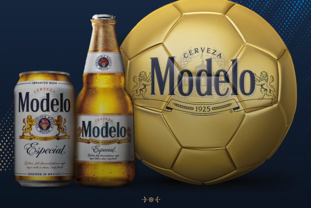 Two bottles of a light-colored beer are positioned beside a can, with a large golden soccer ball displaying an emblem. The background is dark blue, enhancing the vibrant colors of the setup, featured in the Modelo 2026 Soccer Sweepstakes