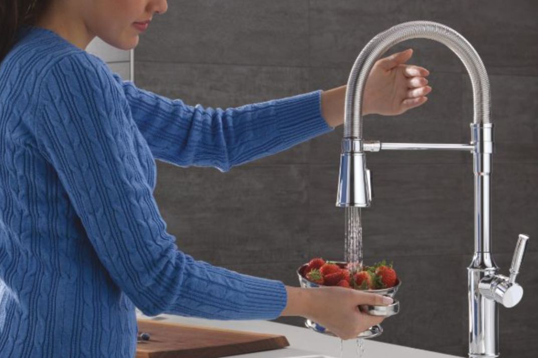 A person in a blue sweater rinses strawberries in a metal bowl under a modern kitchen faucet, with water flowing down and a wooden cutting board in the background, featured in the Delta $3,500 Kitchen Upgrade Sweepstakes