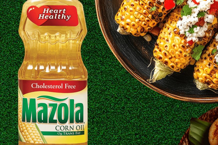 A bottle of corn oil sits beside a plate of grilled corn on the cob topped with cheese, herbs, and red peppers, with a green background adding freshness to the scene, featured in the Mazola Cooler Sweepstakes