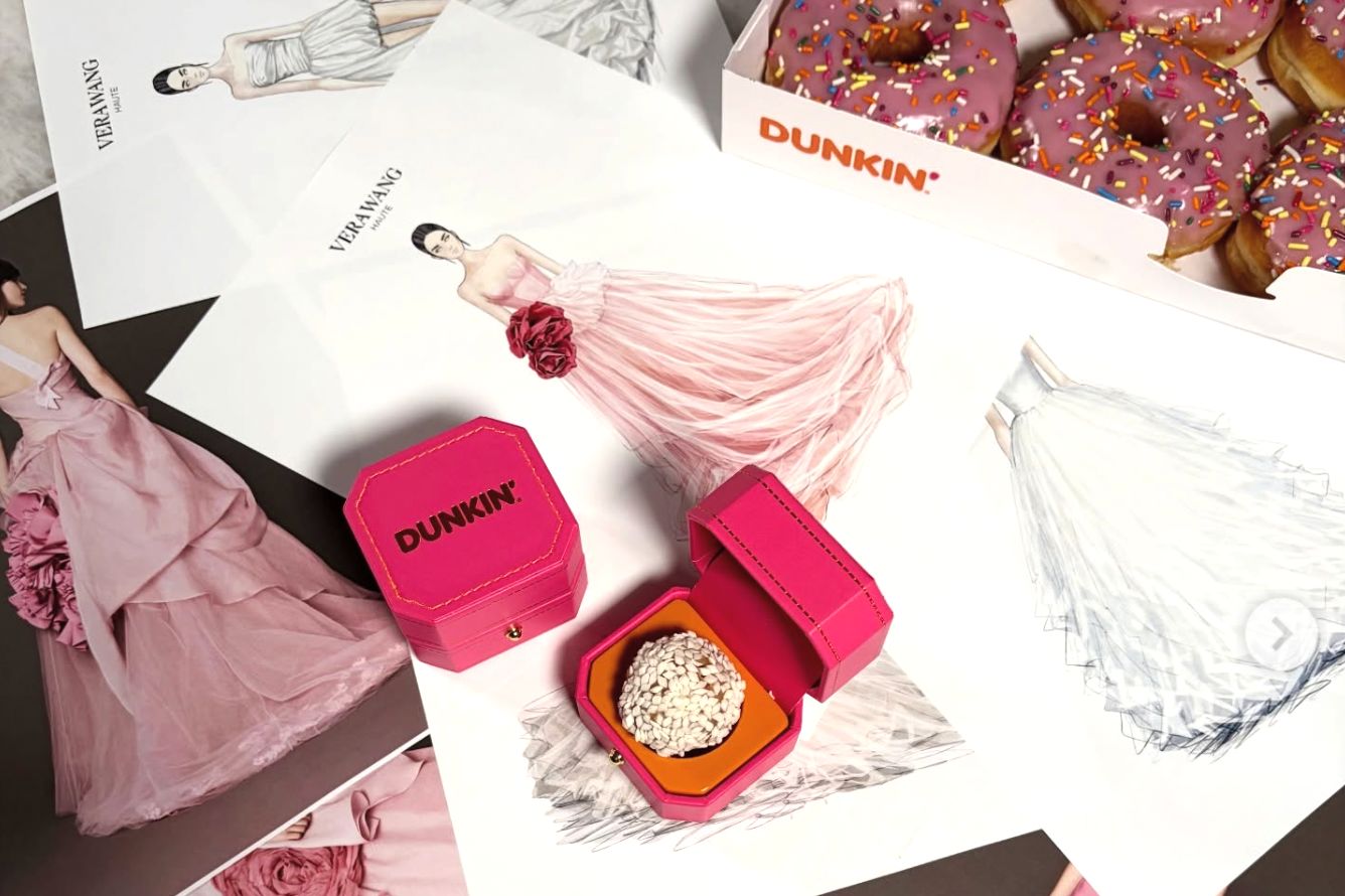 A vibrant pink box sits open revealing a treat, surrounded by fashion sketches of elegant gowns and a box of sprinkled donuts, creating a playful blend of style and indulgence, featured in the Dunkin Vera Wang Sweepstakes