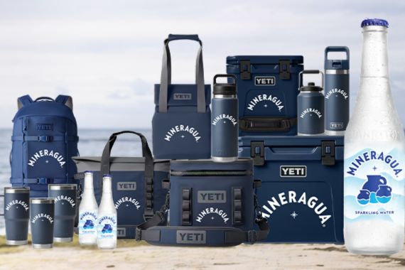 A collection of outdoor gear and drinkware arranged on a sandy beach, featuring coolers, bags, bottles, and cups, showcasing a blue and white color scheme with a sparkling water bottle prominently displayed, featured in the Novamex Yeti Bundle Sweepstakes
