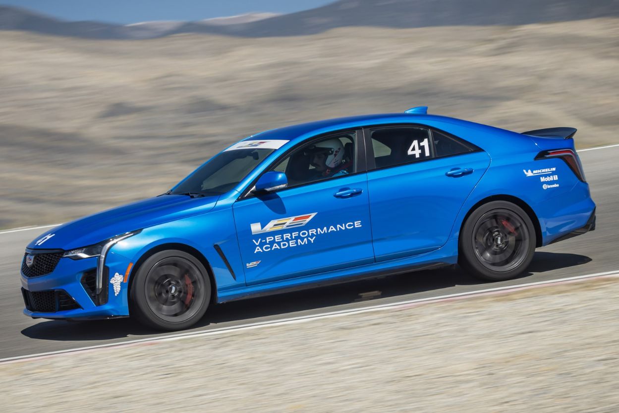 A blue Cadillac racing car speeds around a track, showcasing dynamic movement and performance, with a driver focused on navigating the course. The landscape features a dramatic, blurred backdrop, featured in the Cadillac V-Performance Academy Sweepstakes
