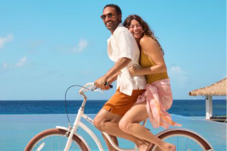 A man and woman enjoy a bike ride together by the pool, laughing under a clear blue sky, with the ocean in the background and a tropical setting enhancing the joyful atmosphere, featured in the Vacation Goals in Curaçao Sweepstakes