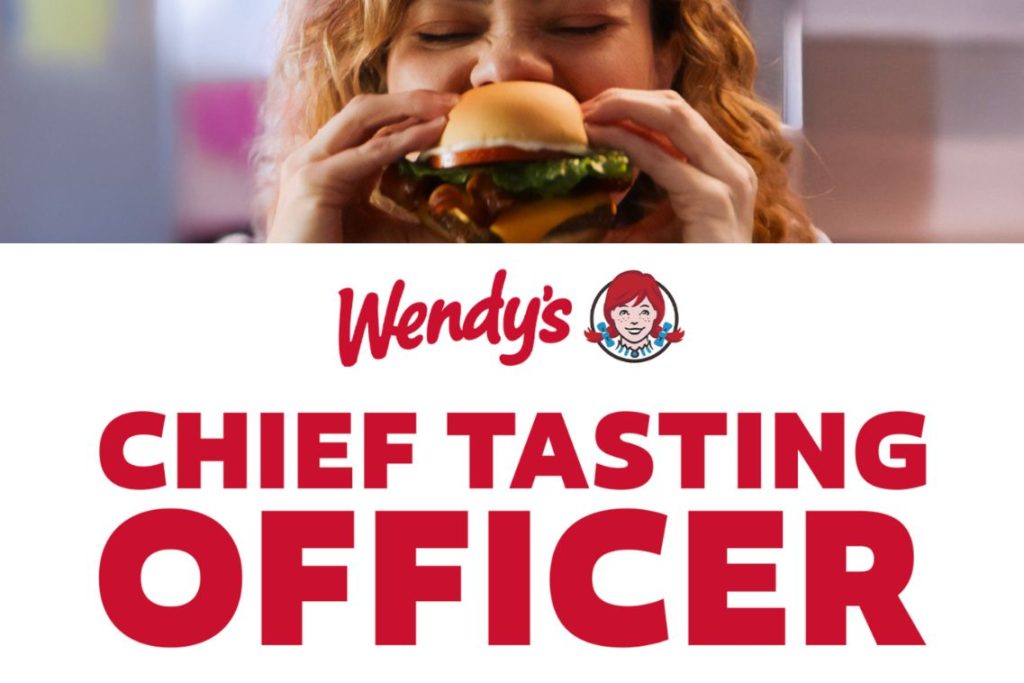 A smiling woman prepares to take a big bite of a hamburger, enjoying a moment of anticipation and excitement in a colorful setting, featured in the Wendy's Chief Tasting Officer Contest