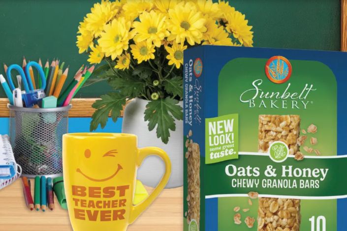 A cheerful yellow mug with a smiley face and "BEST TEACHER EVER" sits next to a box of oatmeal and honey granola bars, alongside a vase of yellow flowers and colored pencils on a wooden desk, featured in the Sunbelt Bakery Spring Classroom Giveaway