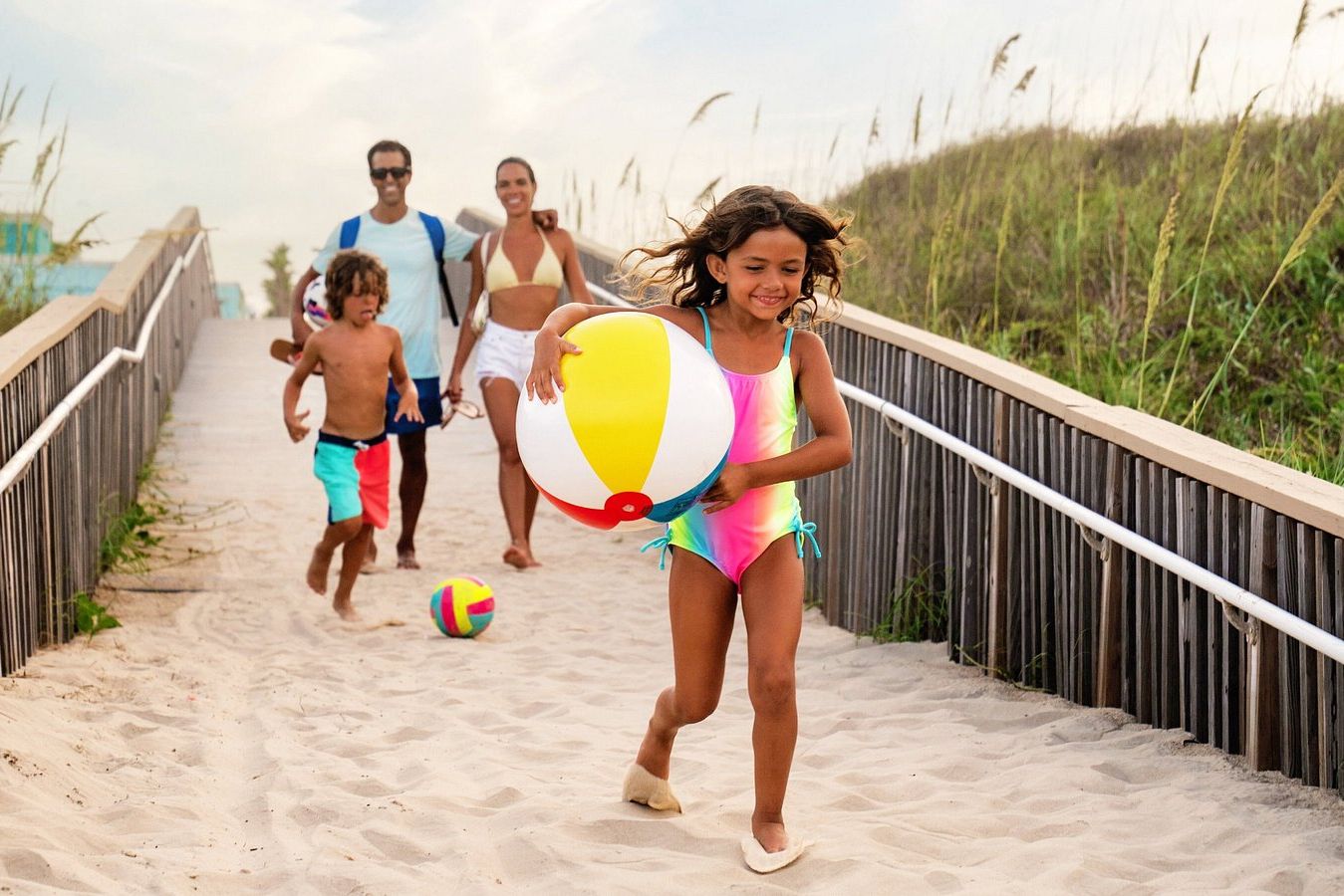 A girl in a colorful swimsuit joyfully runs down a sandy path, holding a beach ball. Two children and adults follow her, enjoying a fun day at the beach, featured in the Tripadvisor South Padre Island Sweepstakes