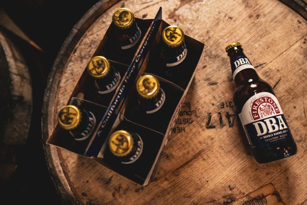 A six-pack of beer bottles sits on a wooden barrel, with one bottle placed beside it. The scene highlights the rich, rustic ambiance of a brewery environment, featured in the Firestone Walker Yeti Cooler Sweepstakes