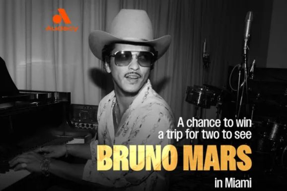 A musician wearing sunglasses and a cowboy hat plays the piano, smiling warmly. The background features soft curtains, adding to the cozy atmosphere, featured in the Bruno Mars Download and Listen