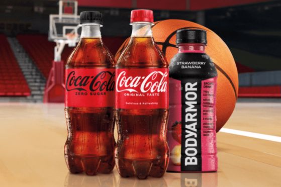 Three bottles of Coca-Cola and a BodyArmor drink are positioned on a basketball court, with a basketball in the foreground and a stadium backdrop, featured in the Coca-Cola Final Four Trip Sweepstakes