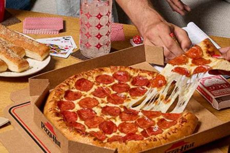 A hand reaches for a slice of pepperoni pizza, with gooey cheese stretching, while another hand holds a drink next to a plate of breadsticks, all set on a wooden table, featured in the Pizza Hut Free Pizza for a Year Sweepstakes