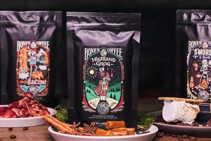 A display of three coffee bags featuring playful, colorful designs, with a focus on the "Highland Grog" blend. Surrounding the bags are spices and snacks, creating a cozy atmosphere, featured in the Bones Coffee Free Coffee Sweepstakes