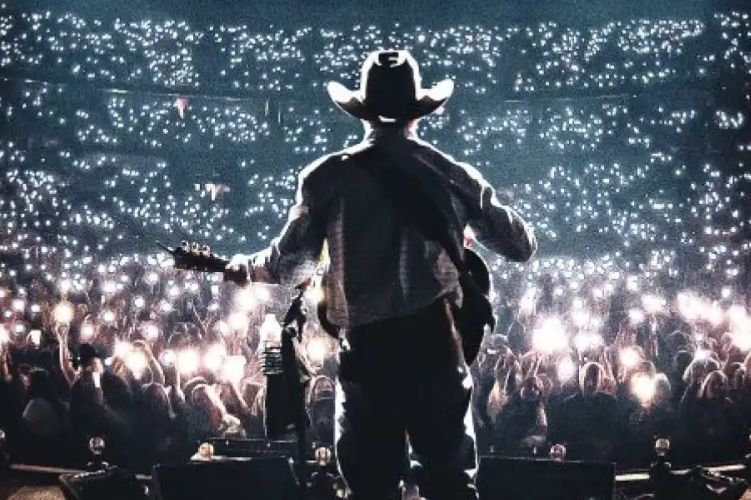 A musician in a cowboy hat plays guitar on stage, facing a sea of concertgoers holding up their phones, creating a shimmering backdrop of lights in a large venue, featured in the George Strait x Cody Johnson Flyaway Sweepstakes