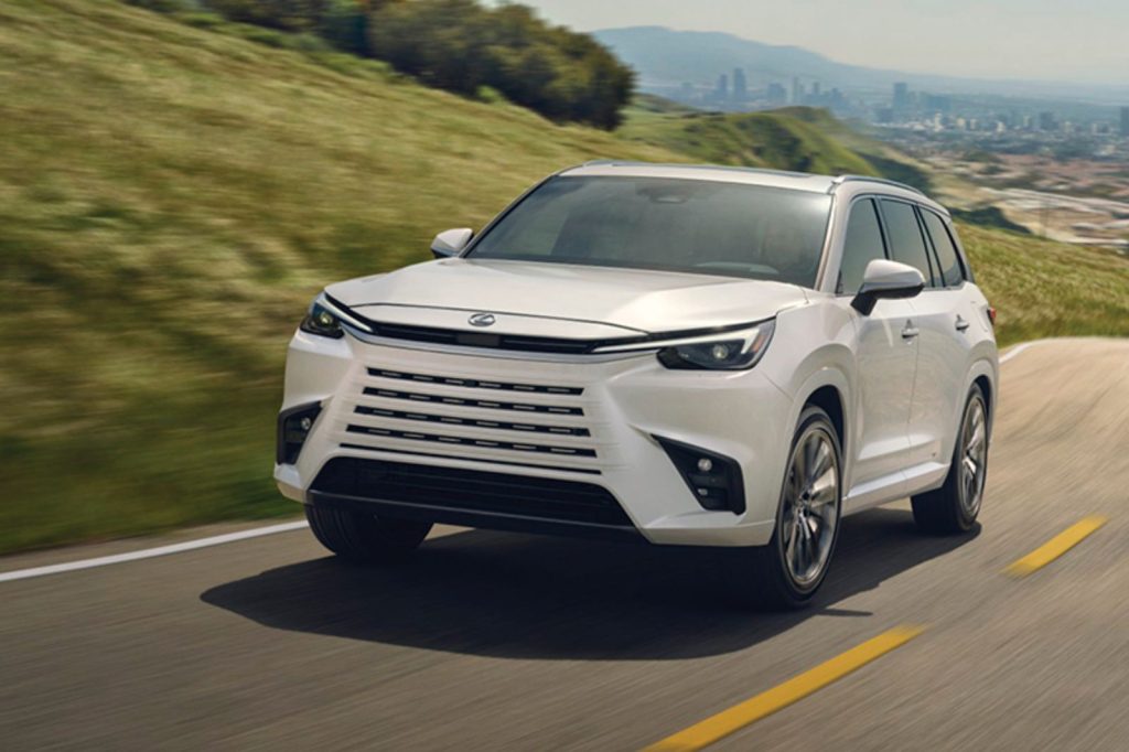 A sleek white SUV drives along a winding road, surrounded by lush green hills and a distant city skyline, showcasing modern design and performance on a sunny day, featured in the Lexus TX Lease Sweepstakes