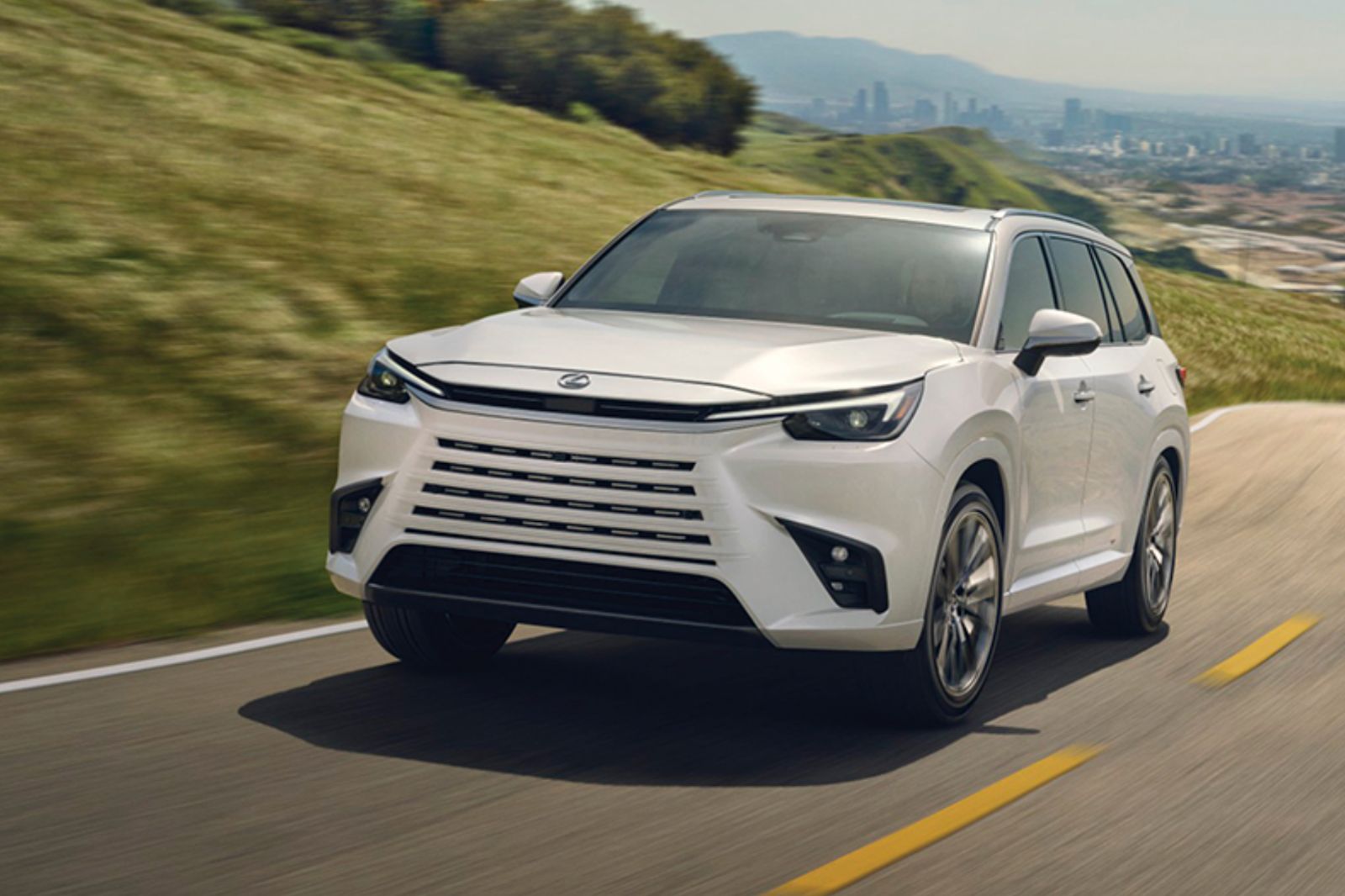 A sleek white SUV drives along a winding road, surrounded by lush green hills and a distant city skyline, showcasing modern design and performance on a sunny day, featured in the Lexus TX Lease Sweepstakes