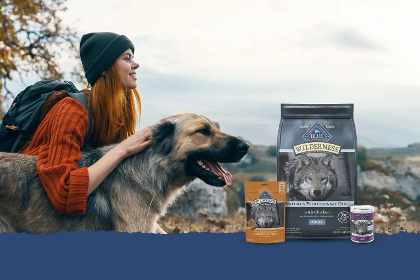 A woman with long red hair and a beanie smiles while petting a happy dog against a scenic outdoor backdrop, with pet food products displayed nearby, featured in the General Mills BLUE Wilderness Sweepstakes