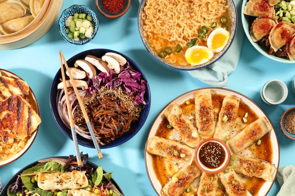 A vibrant spread of Asian cuisine featuring dumplings, noodles, and colorful vegetables, artfully arranged on a blue background with chopsticks and small bowls of toppings, featured in the Pulmuone Lunar New Year Giveaway