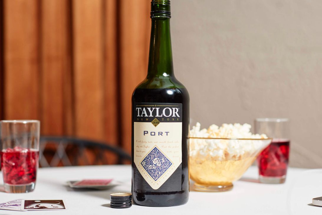 A bottle of port wine is centered on a table, surrounded by glasses of red drinks, a bowl of popcorn, and playing cards, creating a cozy and social atmosphere, featured in the Taylor Port Tastemakers Sweepstakes