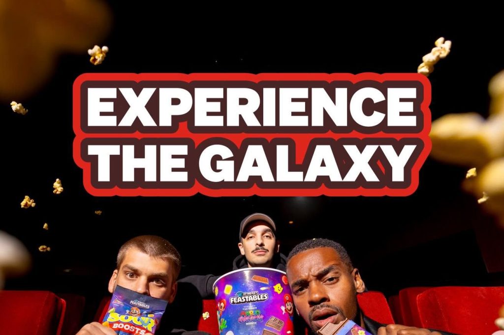 Three individuals pose in a movie theater, holding colorful snacks and looking excited. Popcorn is flying in the air, adding to the lively atmosphere of movie enjoyment, featured in the Feastables Super Mario Galaxy Movie Sweepstakes