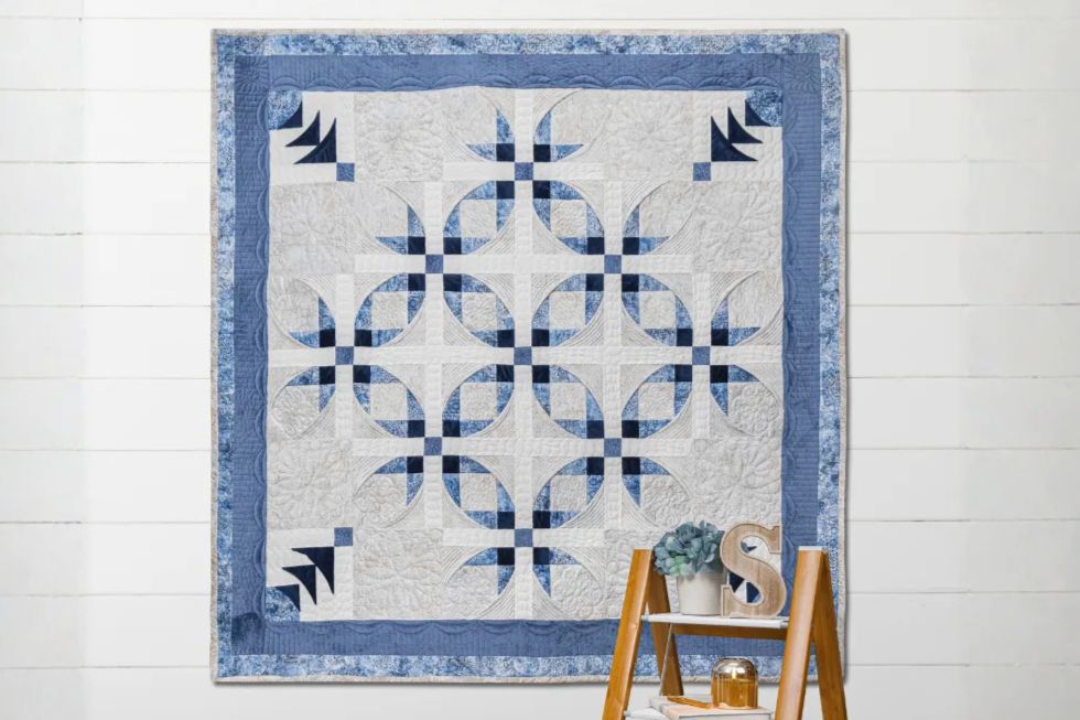 A beautifully crafted quilt featuring a floral pattern in shades of blue and white hangs on a white wooden wall, with a small shelf below holding a potted plant and decorative items, featured in the Shannon Fabrics Sewing Machine Sweepstakes