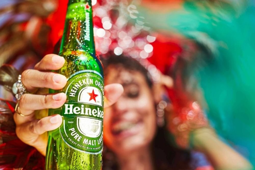 A person joyfully holds a chilled green bottle, splashed with condensation, in a vibrant, festive setting filled with colorful decorations and a joyful atmosphere, featured in the Heineken Sports Sweepstakes