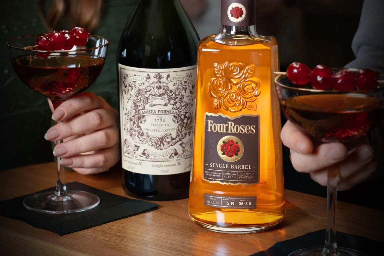 A close-up of two hands holding classic cocktails, one with a garnish of cherries, next to a bottle of bourbon and a bottle of vermouth on a wooden table, featured in the Four Roses Meet Me in Manhattan Sweepstakes