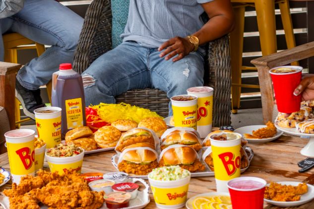 A wooden table is filled with various dishes, including fried chicken, sandwiches, sides, and drinks. People in casual attire relax nearby, enjoying a meal together outdoors, featured in the Bojangles Breakfast for a Year Sweepstakes