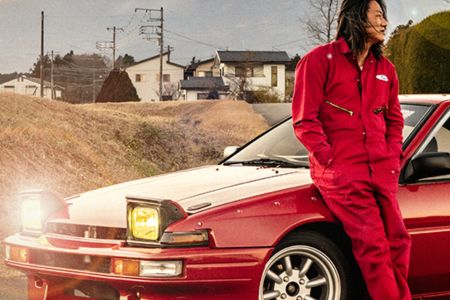 A person in a red jumpsuit leans against a classic red car on a rural road, with houses and trees in the background under a partly cloudy sky, featured in the Horizon Passport Sweepstakes