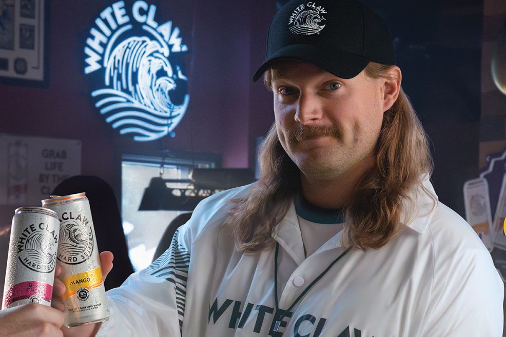 A man with long hair and a mustache poses in a bar, holding two cans of hard seltzer while wearing a baseball-style uniform and cap. A neon logo glows in the background, featured in the White Claw Gift Card Sweepstakes